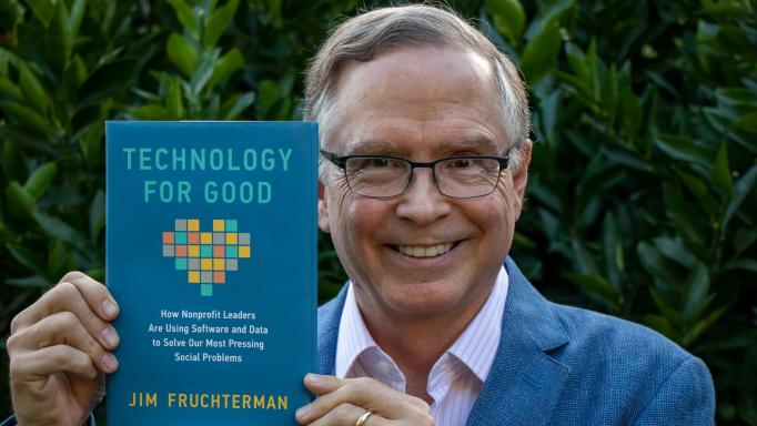 Photo of a smiling Jim Fruchterman holding his recently published book 'Technology for Good: How Nonprofit Leaders are Using Software and Data to Solve Our Most Pressing Social Problems'.  The book has a blue background and a multi-color digitized heart on the front in addition to the title in light blue and white and author's name in yellow.  In the background is a wall of dark green foliage.